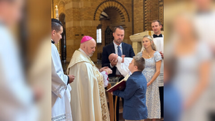 A bishop in ornate robes prays over water during a baptism. Nearby, family members hold a baby for baptism in a church with a brick interior.  A boy in a blue suit holds a prayer book open for the bishop.