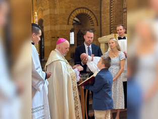 A bishop in ornate robes prays over water during a baptism. Nearby, family members hold a baby for baptism in a church with a brick interior. A boy in a blue suit holds a prayer book open for the bishop.