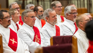 Clergymen in white robes with red stoles sit solemnly in a church during a service.
