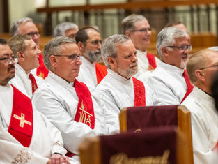Clergymen in white robes with red stoles sit solemnly in a church during a service.