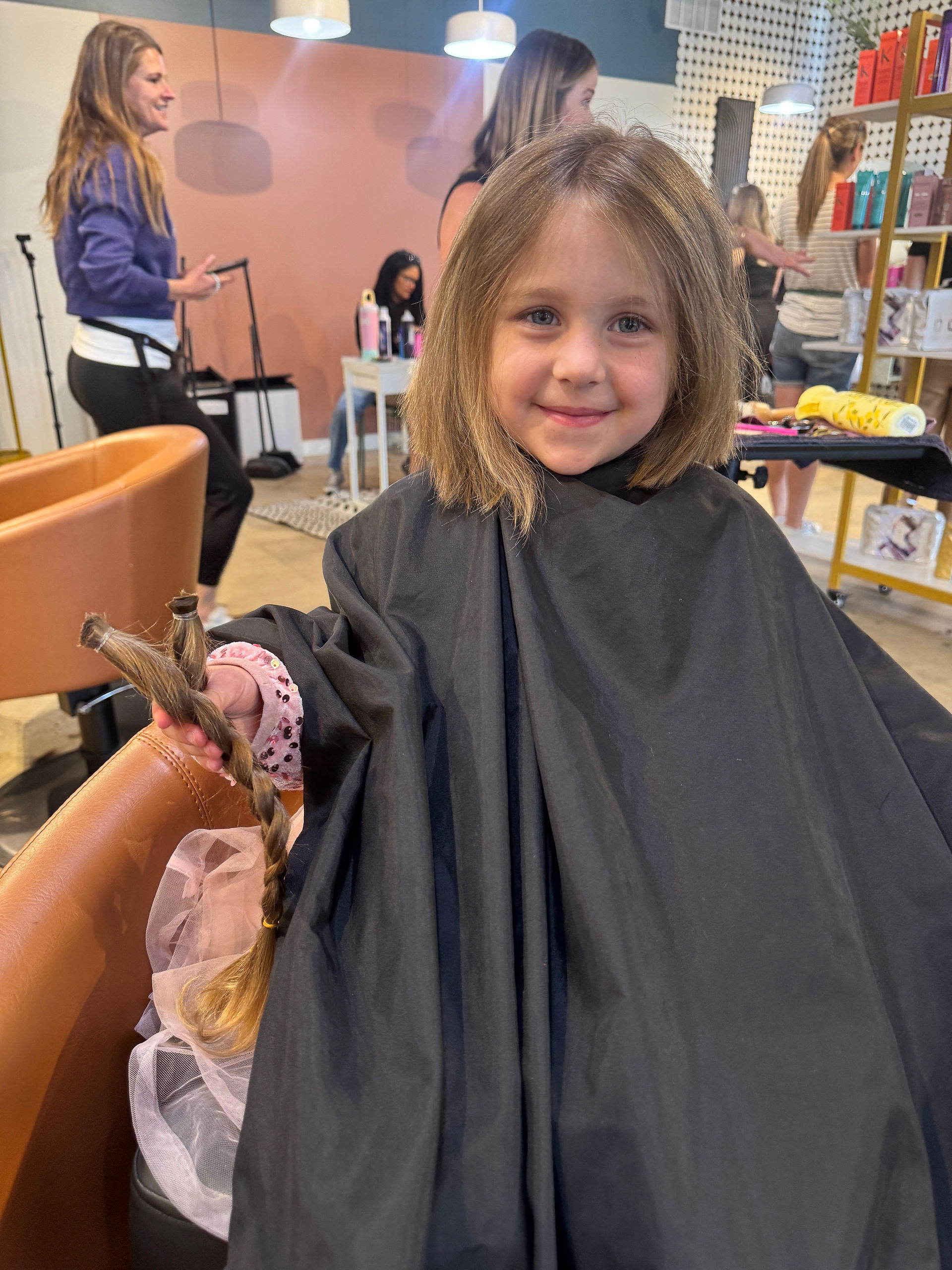 Young girl in a salon chair smiles while holding a freshly cut braid of her hair.