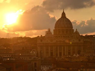 Sunset over St. Peter's Basilica in Rome. The golden sun illuminates the domed structure, with a warm glow casting over the cityscape.