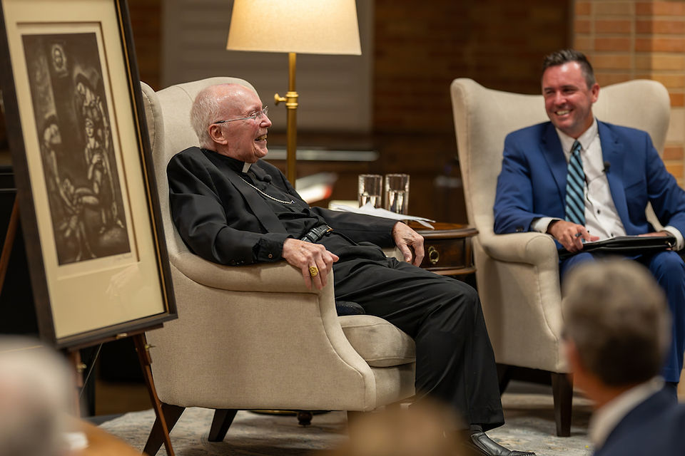 Elderly man in black clerical attire and younger man in a blue suit sit talking in beige armchairs, with a painting and lamp in the background.