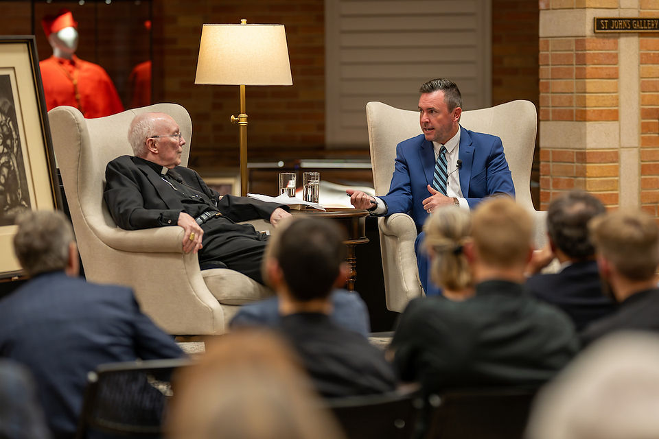 Two men converse in armchairs, one in a black suit, the other in a blue suit. A lamp and red garment in background. Audience visible.