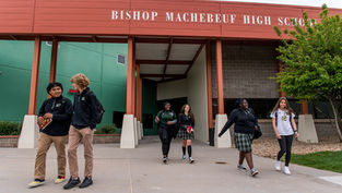 Students walking and talking outside Bishop Machebeuf High School. The setting features green and red school building colors and a tree.