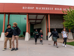 Students walking and talking outside Bishop Machebeuf High School. The setting features green and red school building colors and a tree.