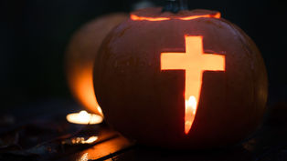 Carved pumpkin with a glowing cross cutout, lit by a candle inside. Dark background and wet surface create a moody, atmospheric scene.