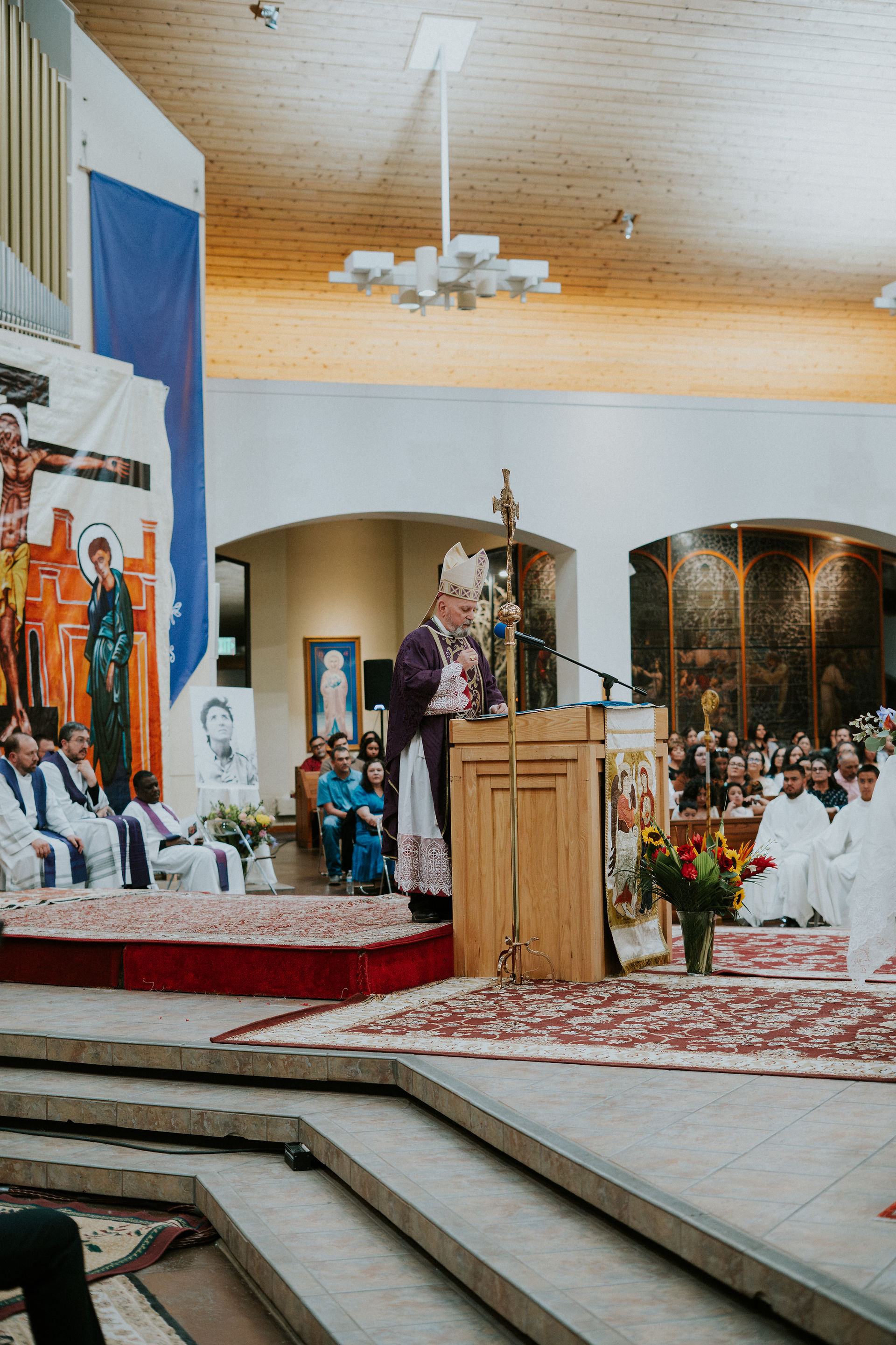 A bishop in ornate vestments speaks at a church pulpit. A congregation listens attentively. Colorful religious artwork adorns the walls.