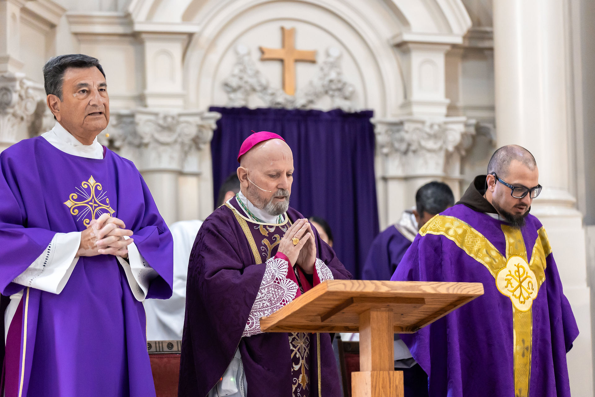 Three clergy members in purple robes stand at an altar, hands clasped in prayer. A church interior with an ornate arch and cross is visible.