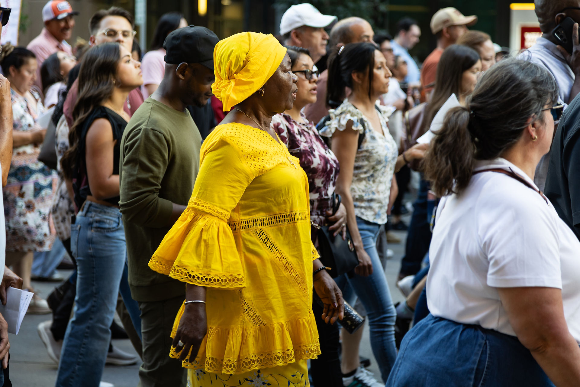 Crowd of people walking at an outdoor event, centered on a woman in a bright yellow dress and headwrap, with focused expressions.
