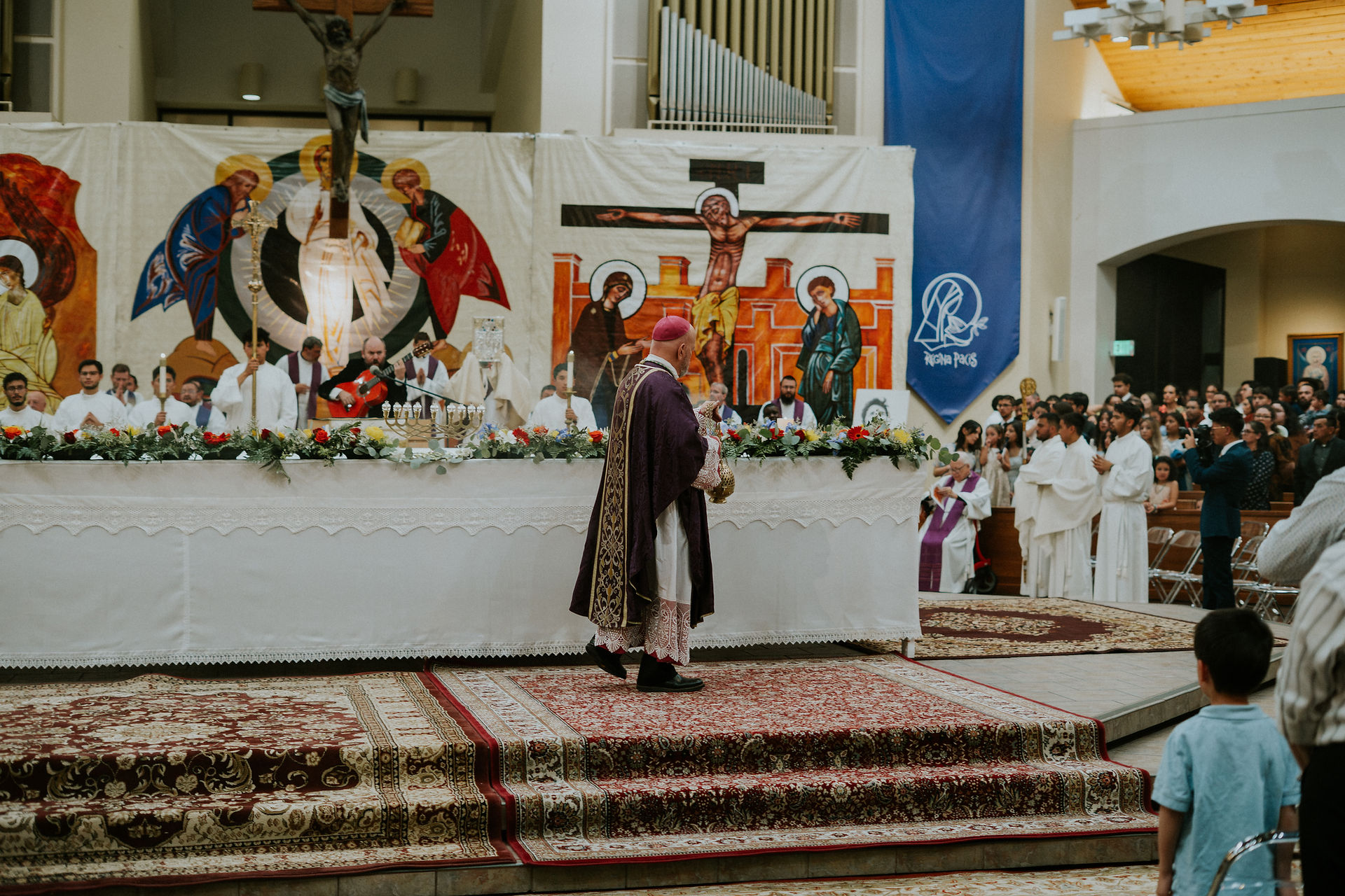 A bishop in purple vestments stands before an altar adorned with flowers. A vibrant religious tapestry and a congregation are in the background.