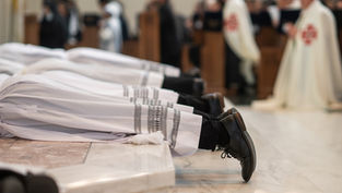 Men in white robes lie prostrate on a marble floor during a religious ceremony, surrounded by blurred figures in formal attire.