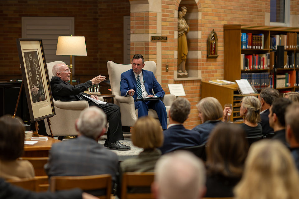 Two men in suits engage in discussion on stage in a library, surrounded by an audience. Brick walls, bookshelves, and statue in background.