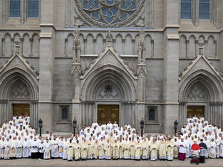 Large group of clergy in white and cream robes pose in front of a grand, ornate cathedral with arched doors, evoking a formal occasion.