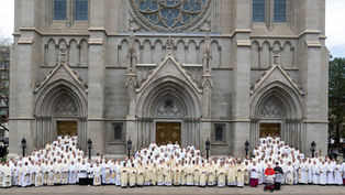 Large group of clergy in white and cream robes pose in front of a grand, ornate cathedral with arched doors, evoking a formal occasion.