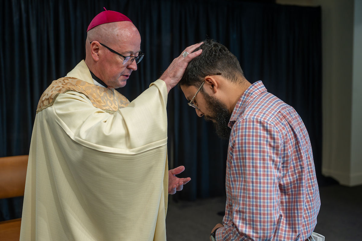 A clergy member in a robe blesses a man in a plaid shirt, pressing his hand on the man's head. Dark curtain backdrop.