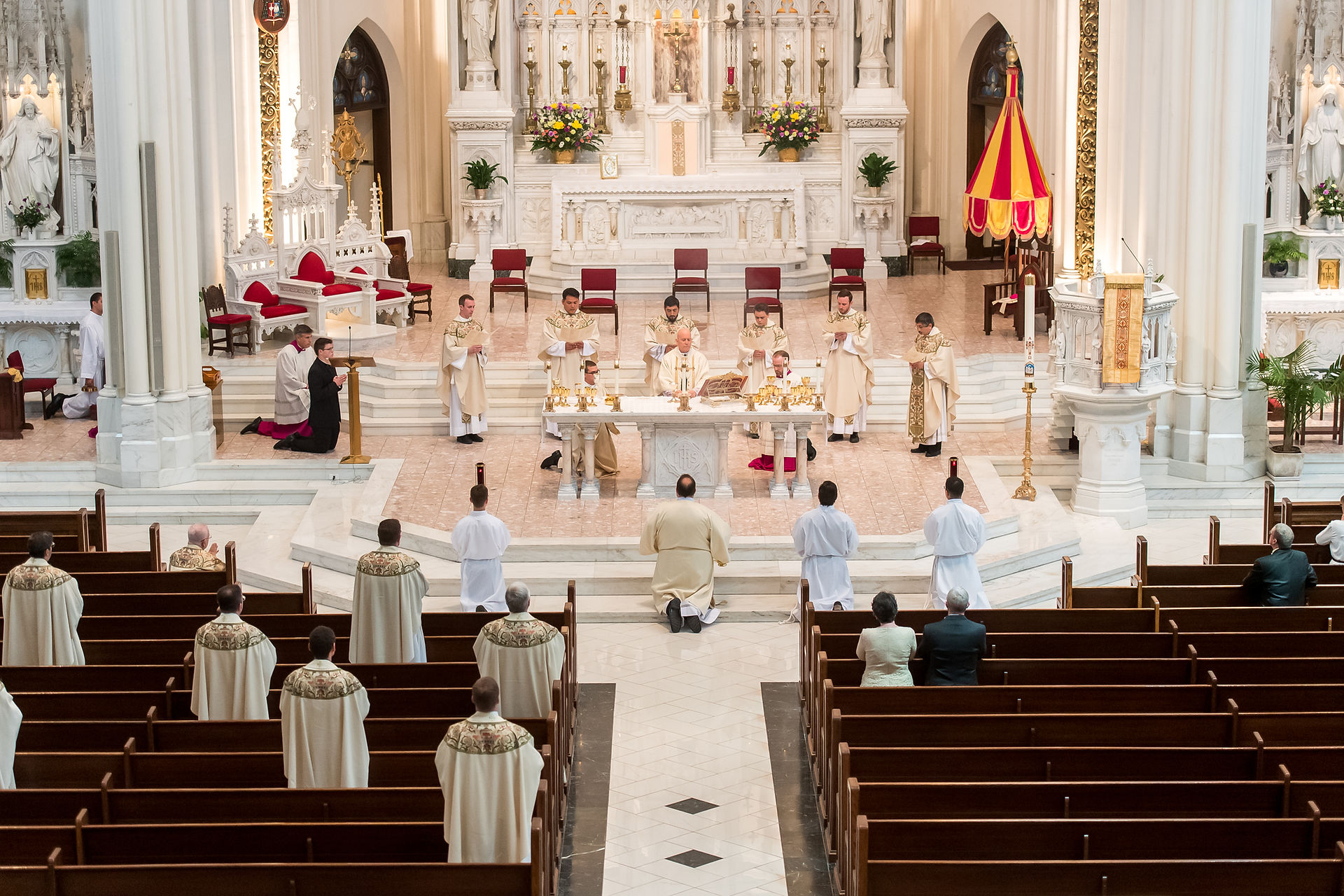 Clergy in beige robes lead a ceremony at a church altar with ornate decorations. Attendees in pews watch solemnly.