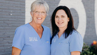 Two smiling women in blue scrubs with "BELLA health + wellness" text. They stand against a gray brick wall with white lettering.