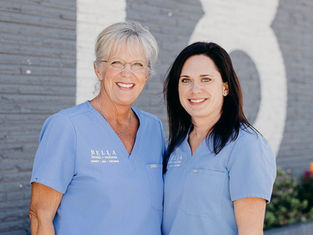 Two smiling women in blue scrubs with "BELLA health + wellness" text. They stand against a gray brick wall with white lettering.