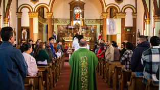 Priest in green robe leads a church procession down the aisle. Congregants stand in pews, ornate altar in background, warm lighting.