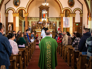 Priest in green robe leads a church procession down the aisle. Congregants stand in pews, ornate altar in background, warm lighting.