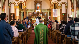 Priest in green robe leads a church procession down the aisle. Congregants stand in pews, ornate altar in background, warm lighting.