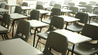 Empty classroom with rows of gray chairs and white desks. Bright windows and beige curtains in the background. Quiet and vacant atmosphere.