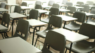 Empty classroom with rows of gray chairs and white desks. Bright windows and beige curtains in the background. Quiet and vacant atmosphere.