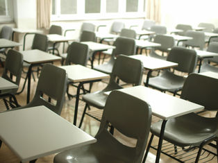 Empty classroom with rows of gray chairs and white desks. Bright windows and beige curtains in the background. Quiet and vacant atmosphere.