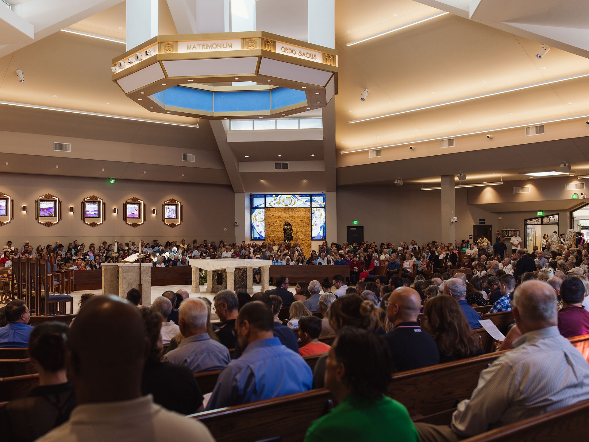 Crowded church interior with people seated and standing. Octagonal light fixture above, stained glass in background, warm lighting.