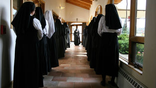 Nuns in black habits line a sunlit corridor with wooden ceiling, either side of a central aisle. A cross is visible on the door.