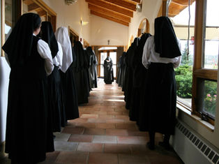 Nuns in black habits line a sunlit corridor with wooden ceiling, either side of a central aisle. A cross is visible on the door.