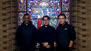 Three men in dark attire stand smiling in front of a vibrant, colorful stained glass window with religious imagery in a brick setting.