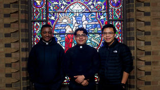 Three men in dark attire stand smiling in front of a vibrant, colorful stained glass window with religious imagery in a brick setting.