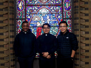 Three men in dark attire stand smiling in front of a vibrant, colorful stained glass window with religious imagery in a brick setting.
