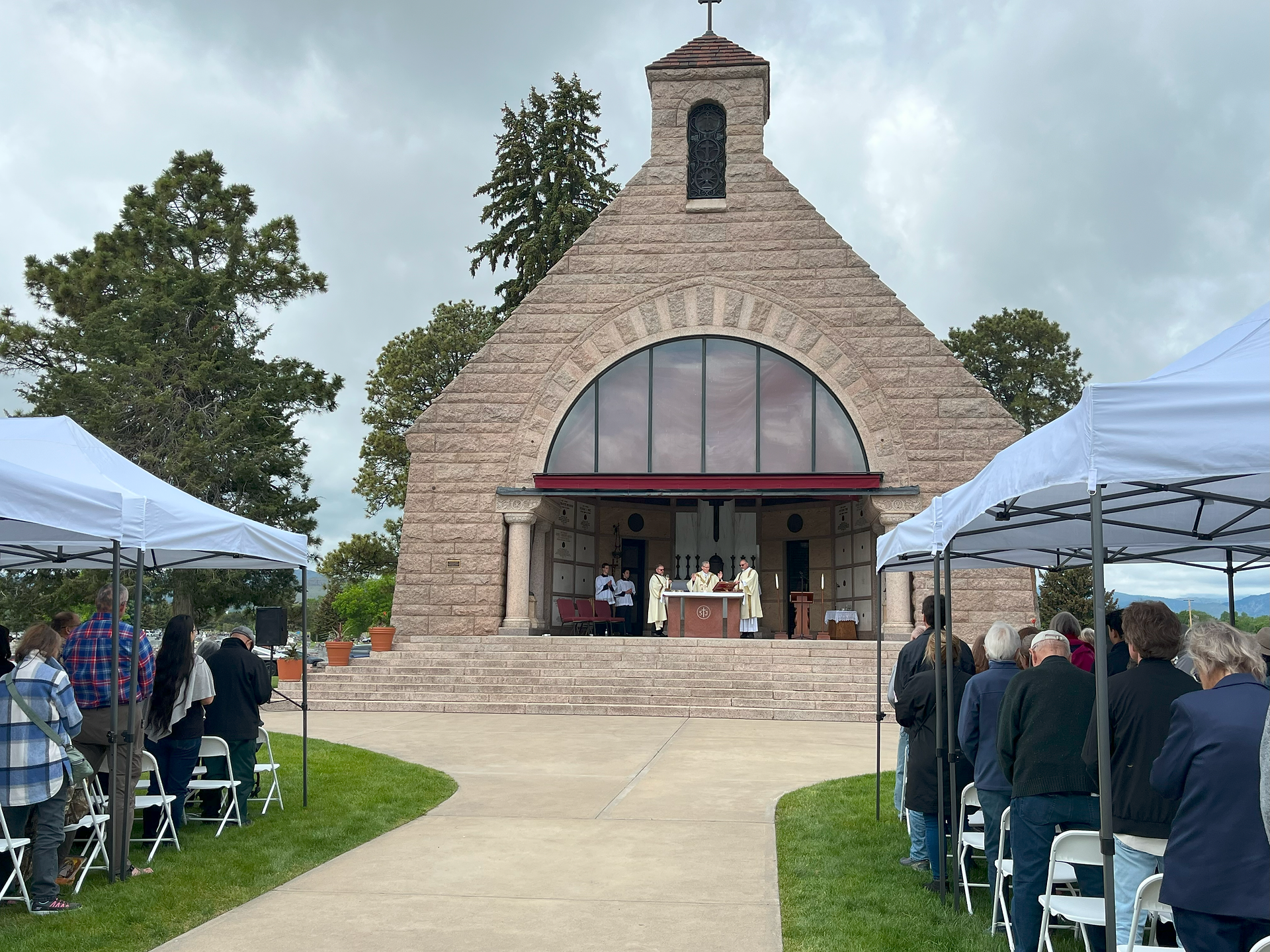 Outdoor church service under gray skies; attendees stand under white tents facing clergy at a stone chapel.