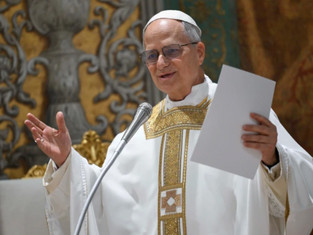 A clergy member in ornate white and gold robes speaks at a microphone, holding papers, with a decorative background. Appears focused.