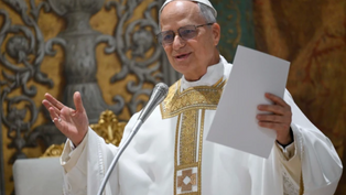 A clergy member in ornate white and gold robes speaks at a microphone, holding papers, with a decorative background. Appears focused.