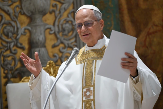 A clergy member in ornate white and gold robes speaks at a microphone, holding papers, with a decorative background. Appears focused.