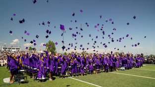Graduates in purple gowns celebrate by tossing caps into the air on a sunny day at a stadium. Crowd watches; mood is joyful and festive.