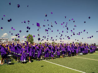 Graduates in purple gowns celebrate by tossing caps into the air on a sunny day at a stadium. Crowd watches; mood is joyful and festive.