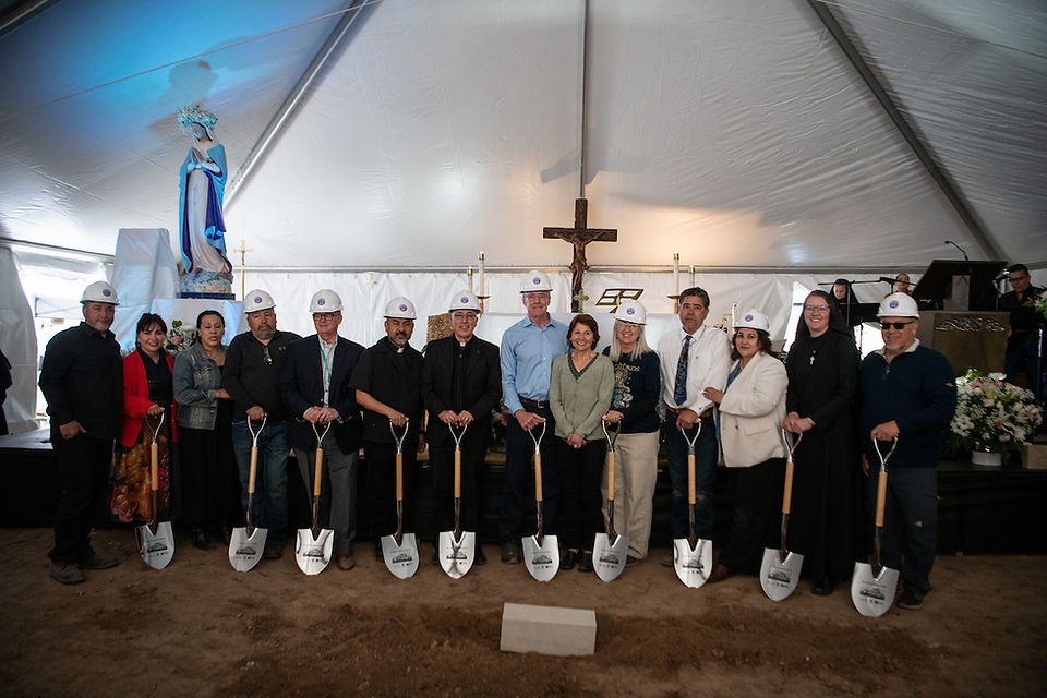 A large group wearing hard hats stands with ceremonial shovels for a commemorative photo inside the event tent.