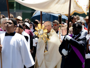 Procession with clergy and participants under a canopy. A priest holds a golden monstrance. Nuns and others follow, some shaded by umbrellas.