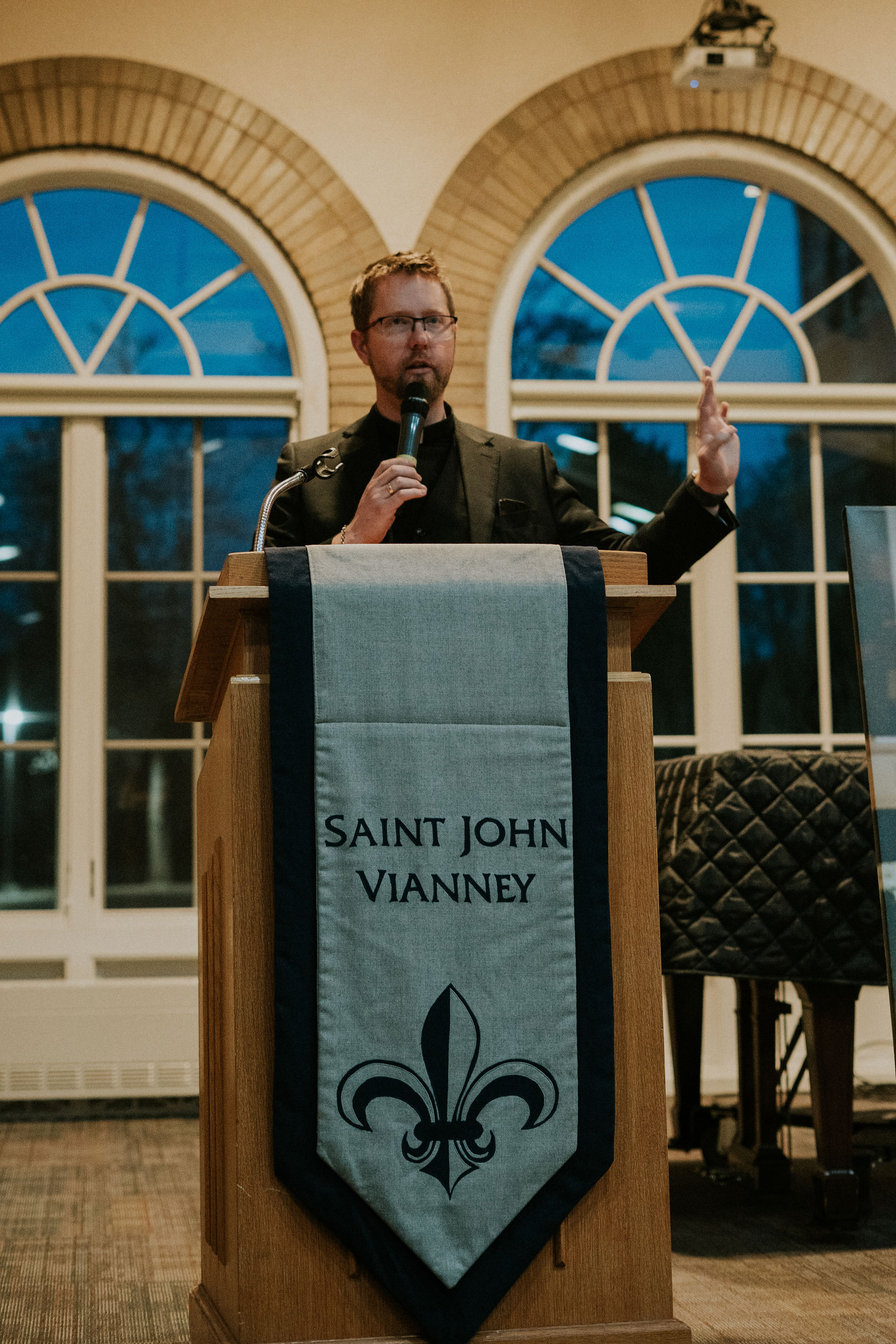 A man in black speaks into a mic behind a podium with "Saint John Vianney" banner. Arched windows and blue evening sky in background.