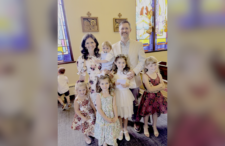 Family posing in church with stained glass windows. Adults hold child, kids in floral dresses smile joyfully. Warm, cheerful atmosphere.
