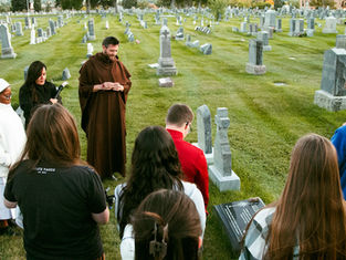 A group gathers in a cemetery, led by a man in a brown robe. A woman carries a candle. The mood is solemn, with green grass and tombstones.