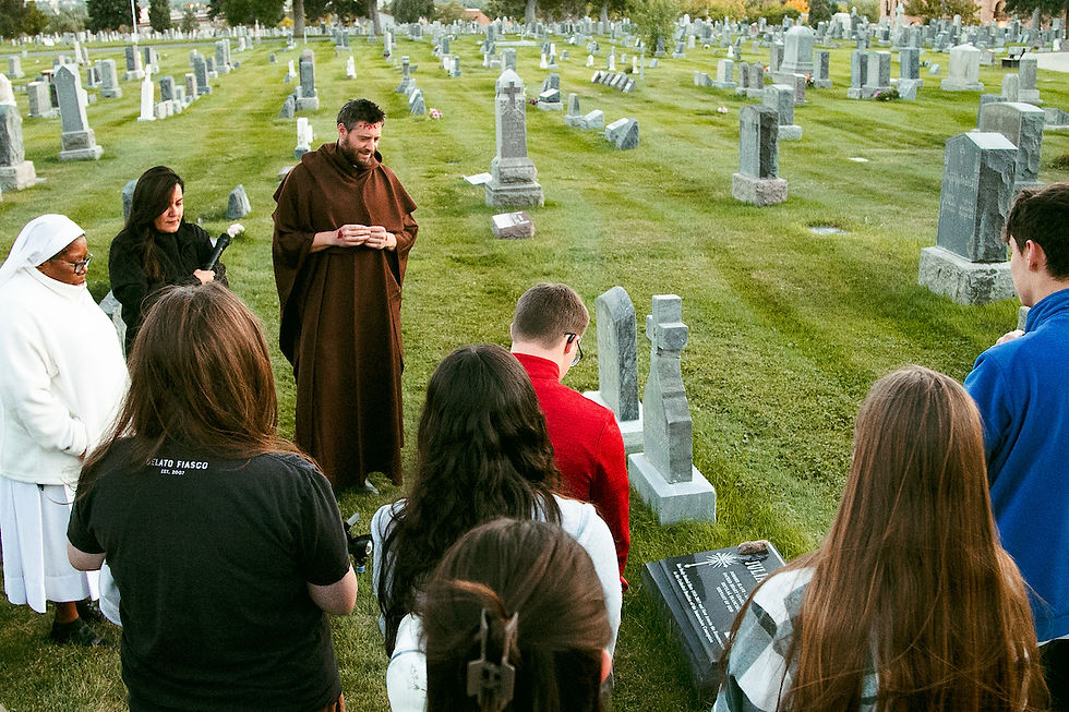 A group gathers in a cemetery, led by a man in a brown robe. A woman carries a candle. The mood is solemn, with green grass and tombstones.