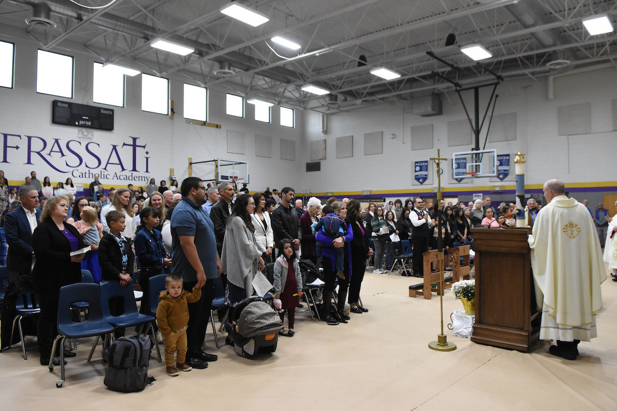 People gathered in a gym for a religious ceremony. A priest stands at a podium. "Frassati Catholic Academy" sign is visible. Calm atmosphere.