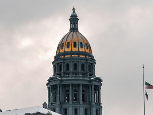 Dome of a classical building with a golden roof against a cloudy sky, American flag flies at half-staff beside it, creating a solemn mood.