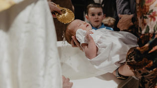 Priest pours water from a gold shell over a sleeping baby in a white gown during a baptism, with family watching nearby.